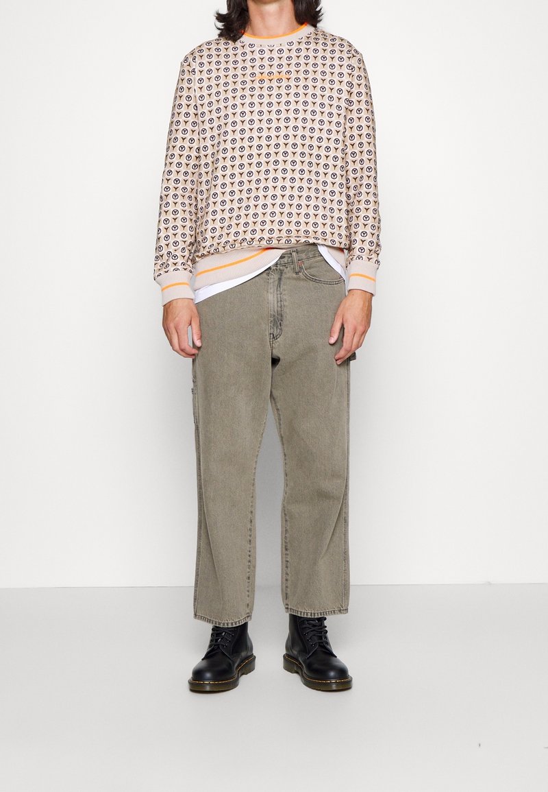 Maglione beige con motivi marroni scuri ripetuti, dettagli arancioni nel colletto e nei polsini. Jeans baggy verde oliva e stivaletti neri con lacci.