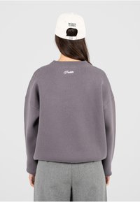 Pull gris surdimensionné en tissu texturé, col rond, avec "Hello" brodé au dos, associé à une casquette beige avec "MODERN YUPPIES."