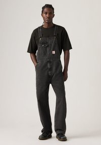 Νεαρός άνδρας με πλεκτά μαλλιά, που φοράει μαύρο φαρδύ t-shirt, μαύρη τζιν σαλοπέτα, με τα χέρια στις τσέπες και μαύρα μποτάκια, στέκεται μπροστά σε απλό φόντο.