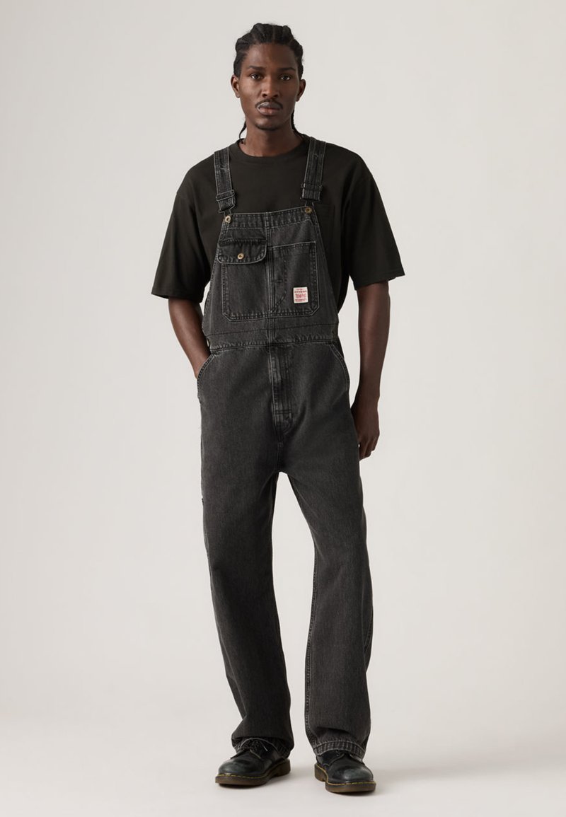 Νεαρός άνδρας με πλεκτά μαλλιά, που φοράει μαύρο φαρδύ t-shirt, μαύρη τζιν σαλοπέτα, με τα χέρια στις τσέπες και μαύρα μποτάκια, στέκεται μπροστά σε απλό φόντο.