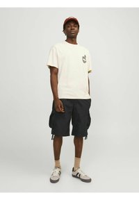 Hellbeige Baumwoll-T-Shirt mit Grafik, schwarze Cargo-Shorts, beige Socken und schwarz-weiße Sneaker. Eine rote Mütze rundet das Outfit ab.