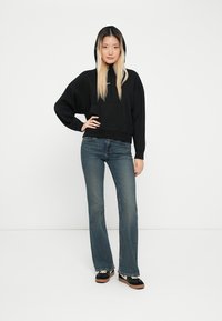 Sweatshirt cropped preto com capucho ajustável, combinado com jeans azuis à boca de sino e sapatos pretos com solas castanhas. Tecido suave, ajuste descontraído.