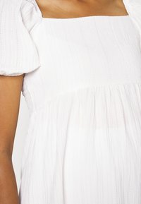 Vestuário branco com decote quadrado, mangas bufantes e tecido texturizado com linhas verticais. Parte inferior evasê com detalhe de design franzido.
