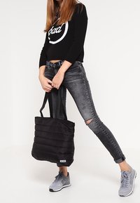 Sac cabas matelassé noir avec texture matelassée et deux poignées en tissu noir. Le modèle porte un jean gris déchiré et des baskets grises.