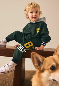 Green sweatshirt and pants set with yellow and white text. White sneakers with Velcro straps. Wooden bench and a corgi in the foreground.
