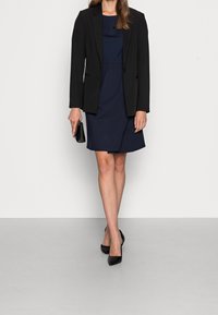 Svart blazer över en marinblå klänning med en figurnära design. Svart kuvertväska i handen och svarta pumps med spetsad tå fulländar outfiten.