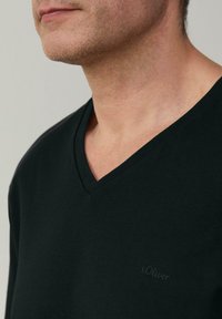 Czarna koszulka z dekoltem w serek, wykonana z bawełny o gładkiej fakturze, z delikatnym logo na klatce piersiowej. Zbliżenie na kołnierz.