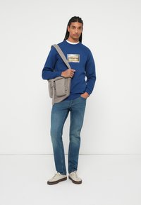Sudadera azul con un gráfico, bolso cruzado gris con cremalleras, jeans azul claro y zapatillas crema con suelas oscuras, sobre un fondo neutro.