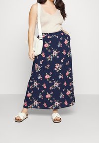 Jupe maxi florale marine avec des motifs floraux roses et verts, associée à un haut beige à col en V et des sandales blanches à glisser. Le sac est tricoté et de couleur crème.