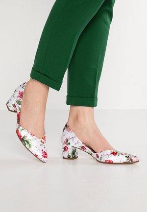 Person iført grønne bukser med opkrogede manchetter og hvide sko med lav blokhæl med blomsterprint, stående på en lys gulv.