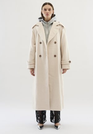 Kermainen kaksirivinen trench-coat, jossa on suuret napit, leveä kaulus ja hartiateipit, käytetty harmaan vetoketjupaidan ja mustien pitsihousujen kanssa.