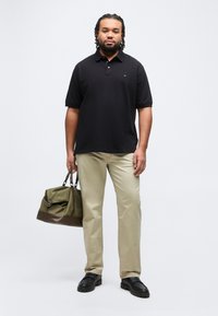 Άνδρας που στέκεται, φορώντας μαύρο πόλο πουκάμισο, μπεζ παντελόνι, μαύρα παπούτσια, κρατώντας μια πράσινη και καφέ τσάντα από τις λαβές, με λευκό φόντο.