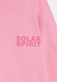 Pinkfarbener Stoff mit dem kräftigen Text "SOLAR SPIRIT", der in einem passenden dunkleren Pink auf der linken Brustseite eines Kleidungsstücks gedruckt ist.