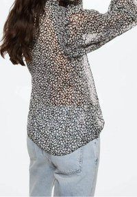 Blouse noire transparente avec un motif floral blanc, col froncé et manches longues. Portée avec un jean en denim délavé, présentant un ourlet incurvé.