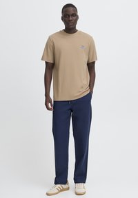 Tricou bej din bumbac cu decolteu rotund și logo mic albastru, asortat cu pantaloni bleumarin și adidași albi cu dungi bej.