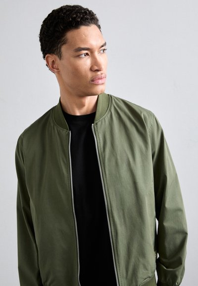 Vihreä pommilentäjä takki, jossa on etuvetoketju, uurretut kaulus ja hihansuut. Kuljetaan mustan t-paidan päällä, ja se on valmistettu sileästä, kevyestä kankaasta.