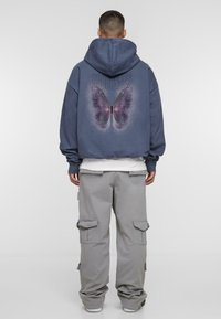 Hooded sweatshirt in dark blue with a large purple butterfly graphic and text on the back; paired with light gray cargo pants.