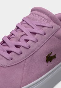 Ροζ σουέντ αθλητικό παπούτσι με υφή, με επίπεδα κορδόνια, χρυσό λογότυπο κροκοδείλου Lacoste και λευκή ελαστική σόλα.