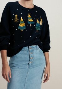 Felpa navy con alberi di Natale ricamati in oro e verde, accentuati da perline bianche. Abbinata a una gonna di jeans azzurro chiaro con bottoni.