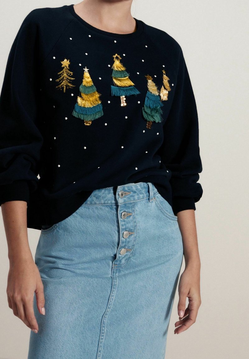 Felpa navy con alberi di Natale ricamati in oro e verde, accentuati da perline bianche. Abbinata a una gonna di jeans azzurro chiaro con bottoni.