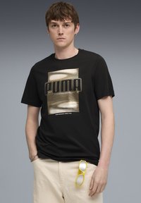 Černé bavlněné tričko s velkým zlatým logem "PUMA" a horizontálními pruhy. Model má na sobě světle béžové kalhoty a žluté sluneční brýle.