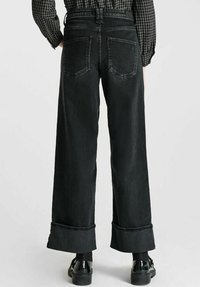Persoon draagt hoogjarige, wijde zwarte jeans met grote manchet, gecombineerd met zwarte schoenen en een geruite overhemd met lange mouwen zichtbaar van achteren.