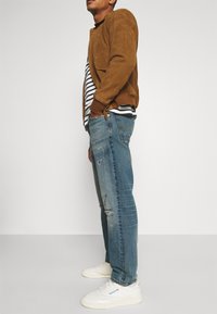 Hnědá semišová bomber bunda, pruhované tričko, opotřebované modré džíny a bílé tenisky. Boční pohled, který ukazuje detaily ležérního outfitu.