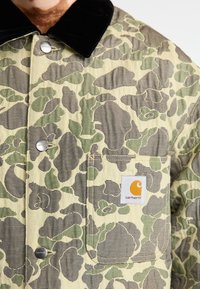 Zunja v bež in zeleni kamuflažni vzorec, z črnim žametnim ovratnikom, srebrnimi dodatki in logotipom Carhartt na žepu.