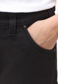 Pantalones de lona negra con un bolsillo frontal, cierre de botón y tejido texturizado. La cinturilla cuenta con presillas para cinturón para mayor soporte.