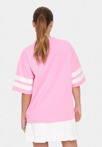 T-shirt rosa oversize con strisce orizzontali bianche sulle maniche. Tessuto di cotone morbido e vestibilità rilassata. Indossato con una gonna plissettata bianca.