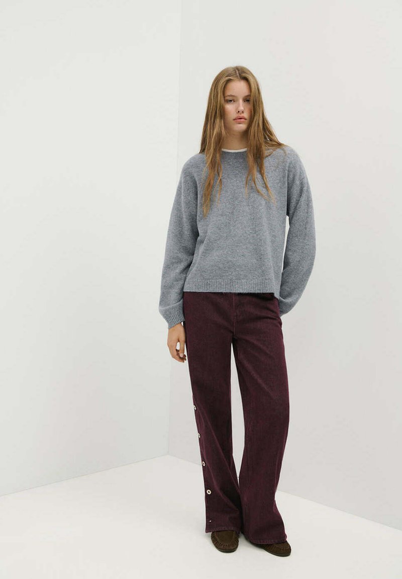 Jeune femme debout portant un pull gris, un pantalon large foncé avec des boutons sur le côté et des chaussures marron sur un fond blanc uni.