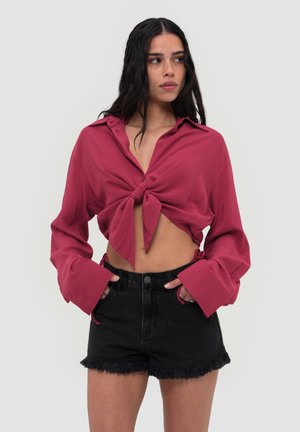 Vrouw met lang donker haar die een geknoopt diep roze overhemd en hoge taille zwarte denim shorts met rafelige randen draagt.