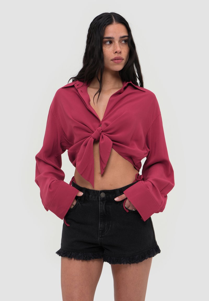 Femme aux longs cheveux foncés portant une chemise rose foncé nouée et un short en jean noir taille haute avec des bords effilochés.