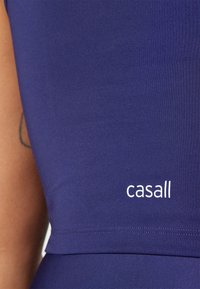 Tamno ljubičasta prilagođena atletska majica od glatkog, rastezljivog materijala, sa belim "casall" logotipom sa leve strane.