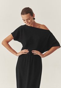 Rochie neagră, cu umerii la vedere, cu mâneci scurte; are un material texturat și o talie elastică cu un accente de nod.