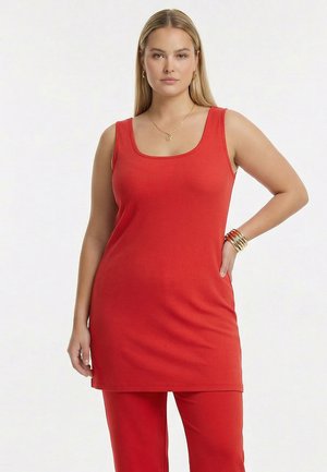 Vrouw met lang blond haar die een mouwloze rode tuniek en bijpassende rode broek draagt, geaccentueerd met gouden sieraden en armbanden.