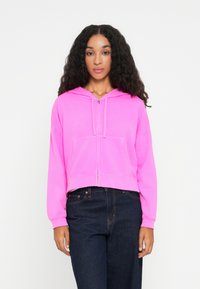 Felpa rosa tagliata con cappuccio, chiusura anteriore con zip e tasca a marsupio; presenta maniche lunghe e una texture morbida e liscia. Abbinata a jeans scuri.