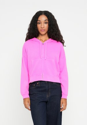 Avkuttet rosa hettegenser med hette, frontlukking med glidelås og lomme; har lange ermer og en myk, glatt tekstur. Kombinert med mørke jeans.