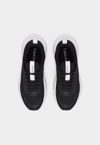 Ζεύγος μαύρων και λευκών αθλητικών παπουτσιών Calvin Klein με μαύρα κορδόνια και ετικέτες της μάρκας στη γλώσσα και στη σόλα, εμφανιζόμενα από πάνω σε λευκό φόντο.