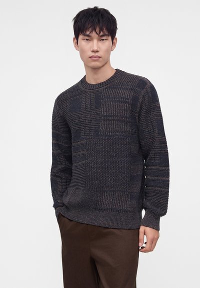 Jeune homme portant un pull en maille texturée sombre et un pantalon marron, debout devant un fond clair uni.