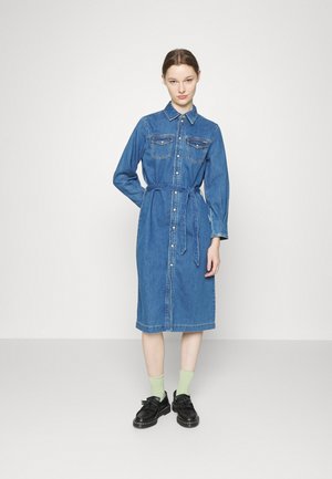 Vestido de camisa em denim, azul claro, com mangas longas, fecho de botão à frente, dois bolsos no peito, cintura cinturada e barra até ao tornozelo. Sapatos pretos e meias verdes.