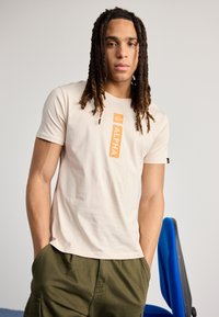 Creme kortærmet t-shirt med en orange "ALPHA" grafisk tryk på fronten. Materialet er blødt bomuldsstof, med en afslappet pasform.