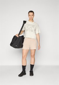 Hellgraues, kürzer geschnittenes T-Shirt mit Grafikdesign, beigefarbene gerippte Shorts, schwarze Sneaker und schwarze Sporttasche, stehend vor einem neutralen Hintergrund.
