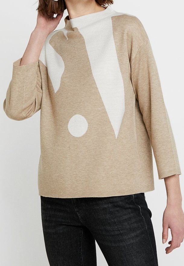 Beige och vit färgblockerad tröja med abstrakta former, buren med svarta jeans, modellens hand rör vid håret nära ansiktet.