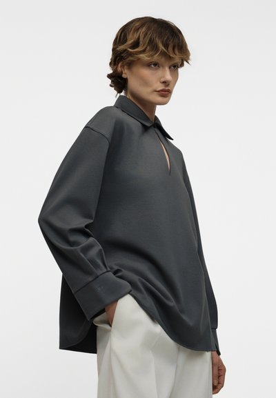 Άτομο με κοντά καστανά μαλλιά που φοράει φαρδιά σκούρα γκρι μπλούζα με μακριά μανίκια και ανοιχτόχρωμο παντελόνι, με τα χέρια μερικώς στις τσέπες.