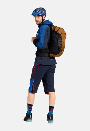 Giacca da ciclismo blu e nera, zaino marrone e nero, pantaloni corti blu con dettagli rossi, calzini blu a fantasia e casco blu.