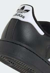 Kruženje crnog Adidas tenisica s petom obloženom bijelim dijelom na kojem se nalazi crni Adidas Trefoil logo i teksturirana potplat.