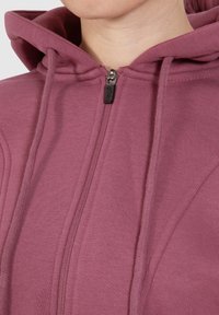 Felpa con cappuccio a zip di colore rosa pallido, in cotone, con zip frontale e texture morbida. Senza motivi o accenti prominenti.