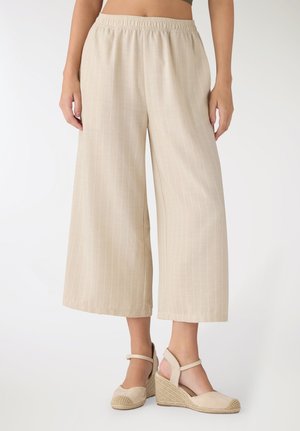 Kobieta ubrana w beżowe, szerokie spodnie 7/8 z elastycznym pasem oraz beżowe koturny espadryle na białym tle.