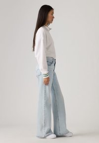 White pullover with a collar and green striped cuffs, paired with light blue wide-leg jeans. Model stands sideways, wearing white shoes.